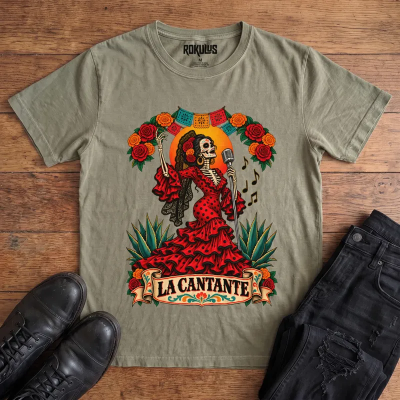 Washed olive Rokulus La Cantante vintage graphic tee laid flat on wood with black boots and jeans, front print showing a Day of the Dead skeleton woman in a red flamenco dress singing into a microphone with roses, marigolds, and papel picado above a “La Cantante” banner