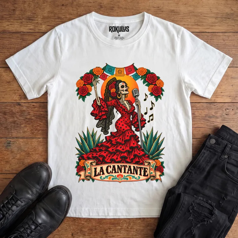 White Rokulus La Cantante vintage graphic tee laid flat on wood with black boots and jeans, front print showing a Day of the Dead skeleton woman in a red flamenco dress singing into a microphone with roses, marigolds, and papel picado above a “La Cantante” banner