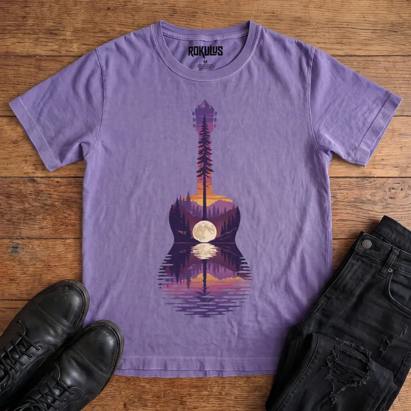 Washed purple Rokulus Guitar Lake vintage graphic tee laid flat on wood with black boots and jeans, front print showing an acoustic guitar formed from a pine forest, mountains, and a full moon reflecting on a lake at sunset in purple and orange