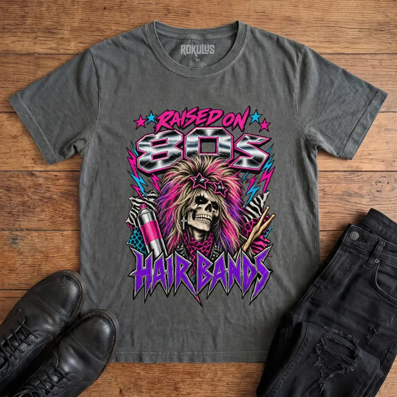 Washed charcoal Rokulus Raised On 80s Hair Bands vintage graphic tee laid flat on wood with black boots and jeans, front print showing a big-haired skeleton glam rocker in star sunglasses with hairspray, drumsticks, and animal print in neon and chrome