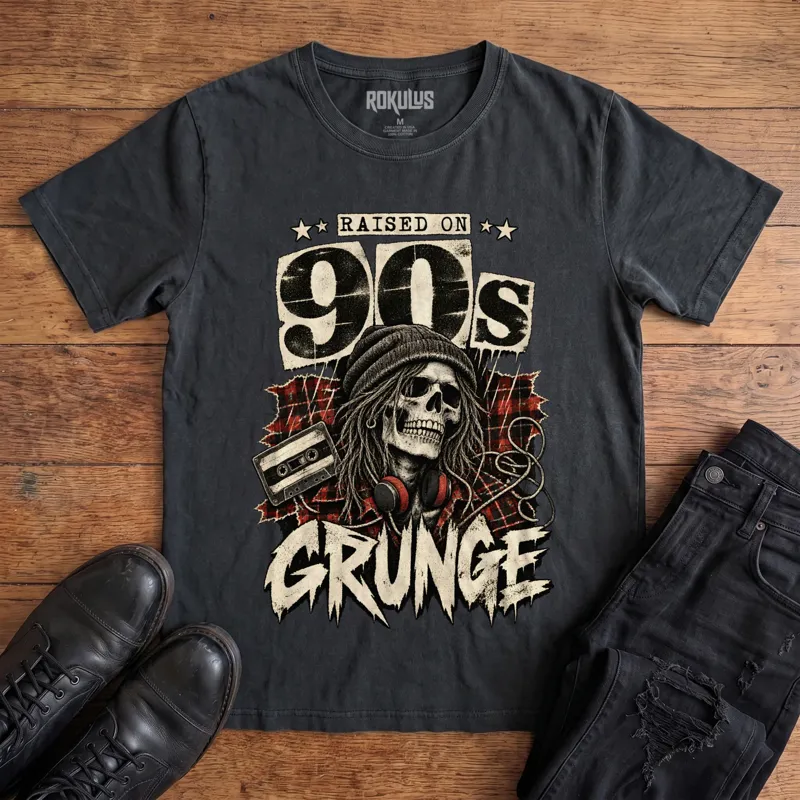 Washed black Rokulus Raised On 90s Grunge vintage graphic tee laid flat on wood with black boots and jeans, front print showing a long-haired skull in a beanie and headphones on red plaid flannel with a cassette tape under “Raised On 90s Grunge” lettering