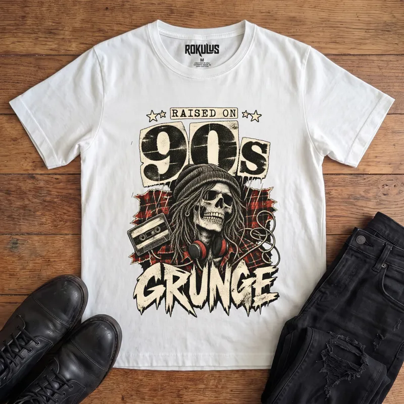 White Rokulus Raised On 90s Grunge vintage graphic tee laid flat on wood with black boots and jeans, front print showing a long-haired skull in a beanie and headphones on red plaid flannel with a cassette tape under “Raised On 90s Grunge” lettering