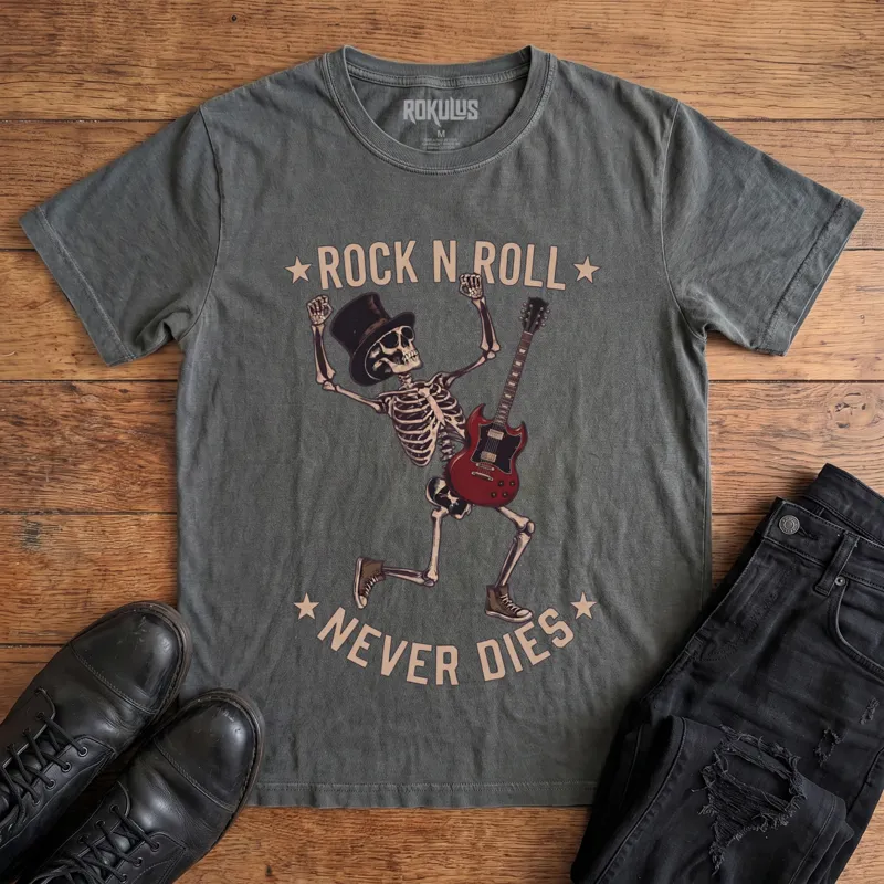 Washed charcoal Rokulus Rock N Roll Never Dies vintage graphic tee laid flat on wood with black boots and jeans, front print showing a dancing top-hat skeleton with a red electric guitar and fists raised under arched "Rock N Roll Never Dies" lettering
