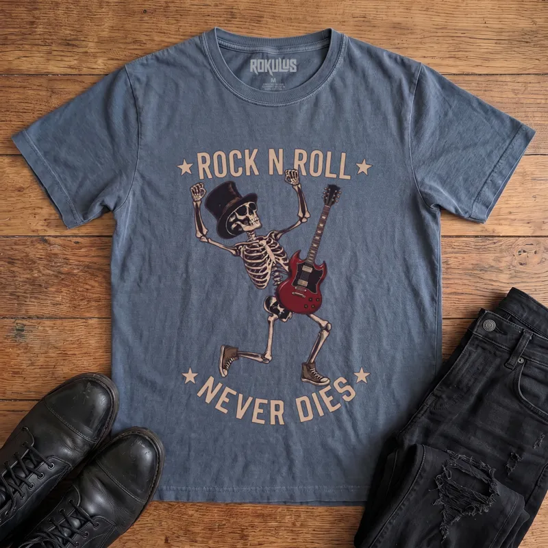 Washed navy Rokulus Rock N Roll Never Dies vintage graphic tee laid flat on wood with black boots and jeans, front print showing a dancing top-hat skeleton with a red electric guitar and fists raised under arched "Rock N Roll Never Dies" lettering