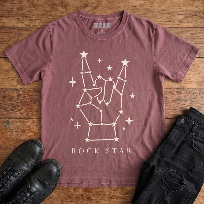 Washed maroon Rokulus Rock Star Constellation vintage graphic tee laid flat on wood with black boots and jeans, front print showing a rock horns hand formed from a star constellation above “Rock Star” lettering in cream