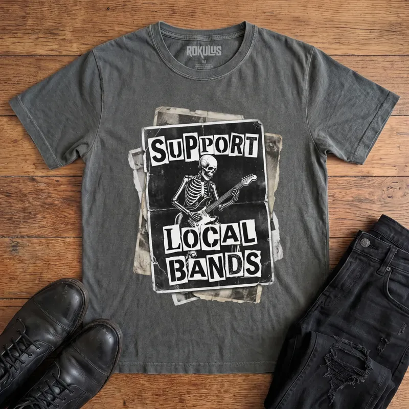 Washed charcoal Rokulus Support Local Bands vintage graphic tee laid flat on wood with black boots and jeans, front print showing a torn punk flyer with a skeleton playing electric guitar and "Support Local Bands" in ransom-note lettering