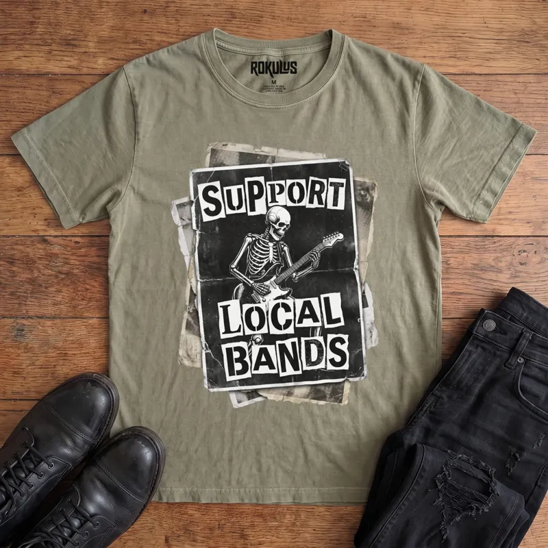 Washed olive Rokulus Support Local Bands vintage graphic tee laid flat on wood with black boots and jeans, front print showing a torn punk flyer with a skeleton playing electric guitar and "Support Local Bands" in ransom-note lettering