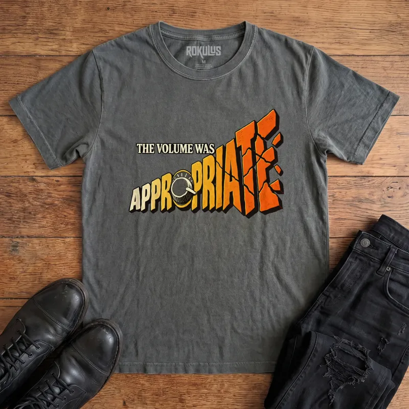 Washed charcoal Rokulus The Volume Was Appropriate vintage graphic tee laid flat on wood with black boots and jeans, front print reading “The Volume Was Appropriate” with a cranked amp knob and shattering orange-to-yellow lettering