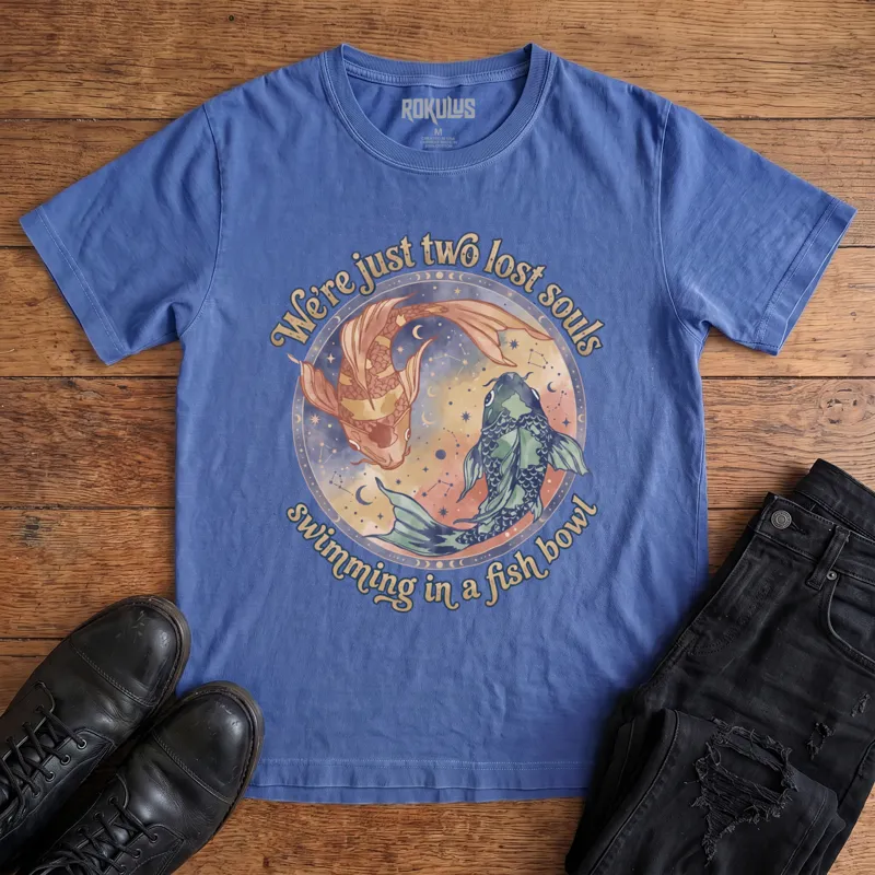 Washed royal blue Rokulus Two Lost Souls vintage graphic tee laid flat on wood with black boots and jeans, front print showing two watercolor koi in a celestial ring of moon phases and constellations with "We're just two lost souls swimming in a fish bowl" in retro script